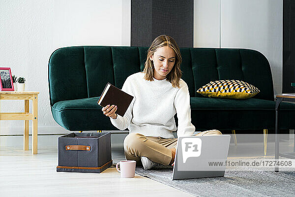 Geschäftsfrau mit Buch  die einen Laptop benutzt  während sie bei einer Kiste auf dem Boden im Büro sitzt