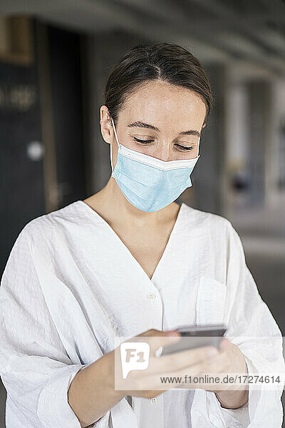 Geschäftsfrau mit Gesichtsmaske  die ein Mobiltelefon benutzt  während sie im Büro steht