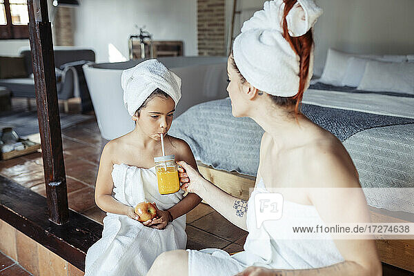 Mother in towel feeding juice to daughter while sitting at home