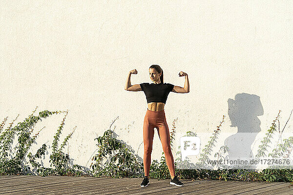 Young female athlete flexing muscle while standing against wall during sunrise
