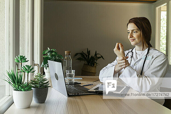 Junge Ärztin  die das Handgelenk hält  während sie während einer Online-Konsultation im Büro zu Hause etwas erklärt