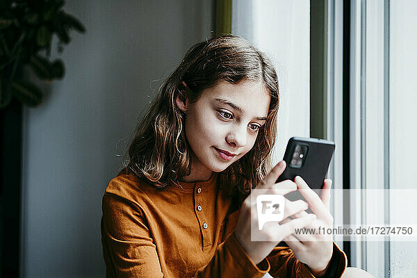Pre-adolescent child using mobile phone while sitting by window at home