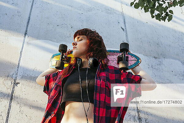Junge rothaarige Frau hält Skateboard hinter dem Kopf mit geschlossenen Augen gegen die Wand