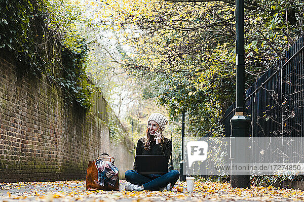 Lächelnde Frau mit Laptop  die auf dem Smartphone spricht  während sie auf der Straße sitzt