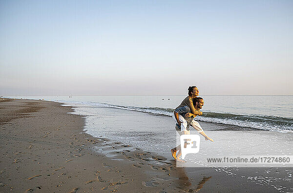 Cheerful man giving piggyback to girlfriend on shore at beach against clear sky during sunset