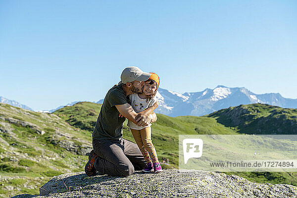 Father kissing daughter while kneeling on rock during sunny day