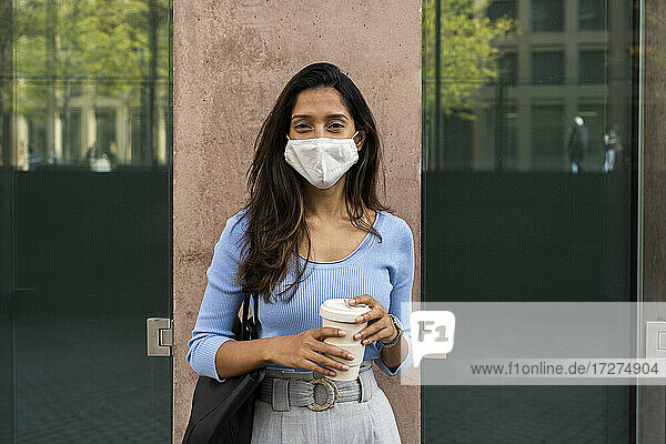 Young businesswoman in protective face mask holding disposable coffee cup while standing against building