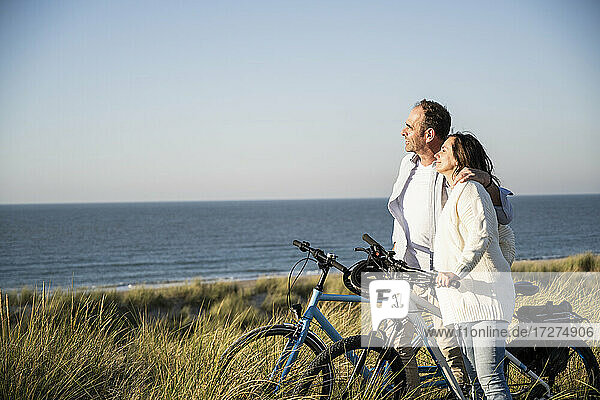Lächelndes Paar mit Fahrrädern  das die Aussicht betrachtet  während es am Strand gegen den klaren Himmel am Wochenende steht