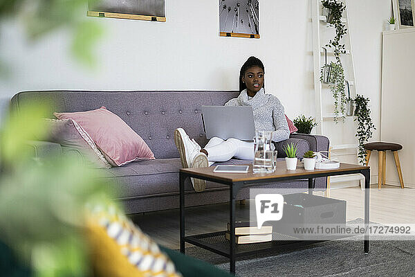 Geschäftsfrau  die wegschaut  während sie einen Laptop benutzt  während sie zu Hause auf dem Sofa sitzt