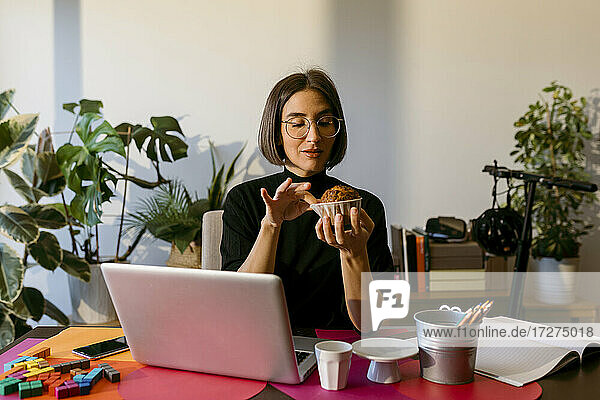 Mid adult businesswoman holding cake while sitting by laptop at home
