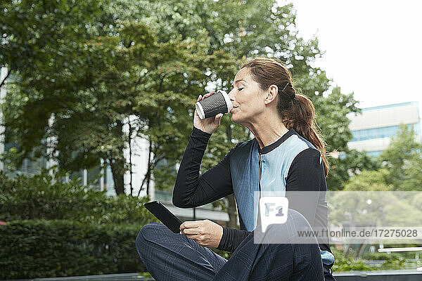 Geschäftsfrau trinkt Kaffee  während sie im Büropark ein Mobiltelefon benutzt