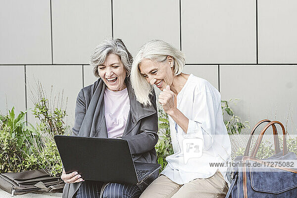 Reife Geschäftsfrau lächelt bei der Arbeit am Laptop vor einem Gebäude