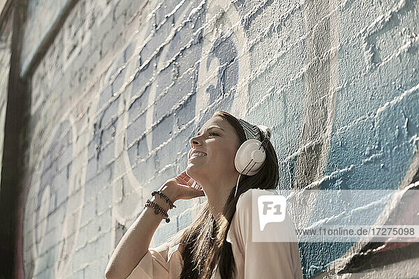 Happy young woman listening music through headphones while standing against wall with graffiti on sunny day