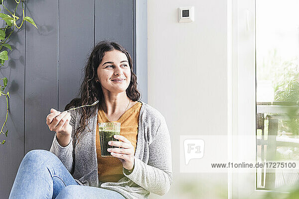 Lächelnde Frau mit Smoothie-Glas  die wegschaut  während sie zu Hause sitzt