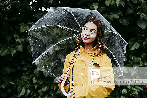 Lächelndes Mädchen im Regenmantel mit Regenschirm  das wegschaut  während es an einer Laubwand steht
