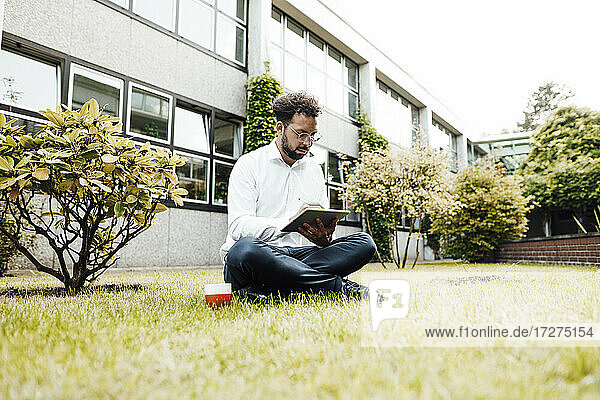 Businessman using digital tablet while sitting with coffee cup on grass against industry