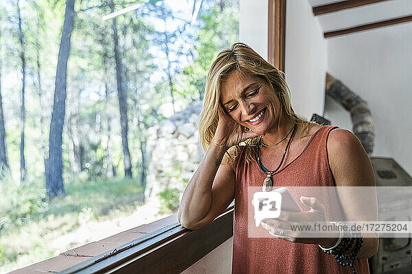 Smiling blond female tourist using smart phone while standing by window at retreat