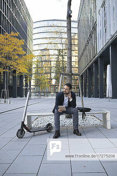 Unternehmer mit digitalem Tablet  der mit einem Elektroroller auf einer Bank in der Stadt sitzt