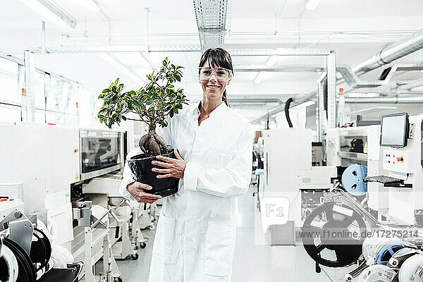 Lächelnde Wissenschaftlerin  die eine Topfpflanze hält  während sie inmitten von Maschinen im Labor steht