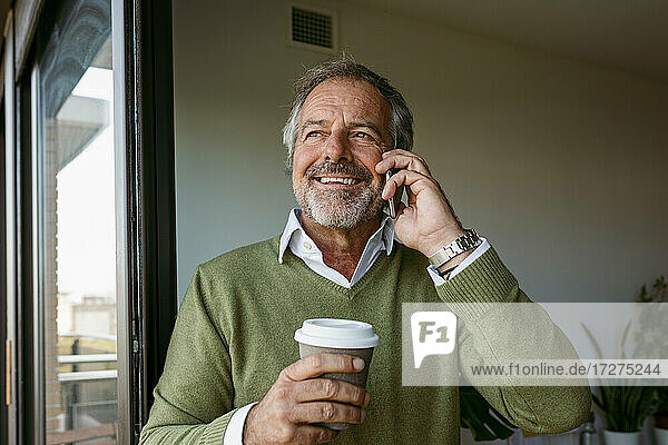 Mann mit Kaffeetasse  der zu Hause steht und mit seinem Handy telefoniert