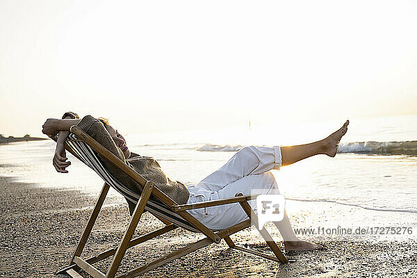 Entspannte junge Frau sitzt auf einem Klappstuhl am Strand bei Sonnenuntergang
