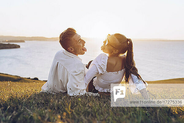 Young couple sticking out tongue while relaxing on hill at Mirador de La Providencia  Gijon  Spain