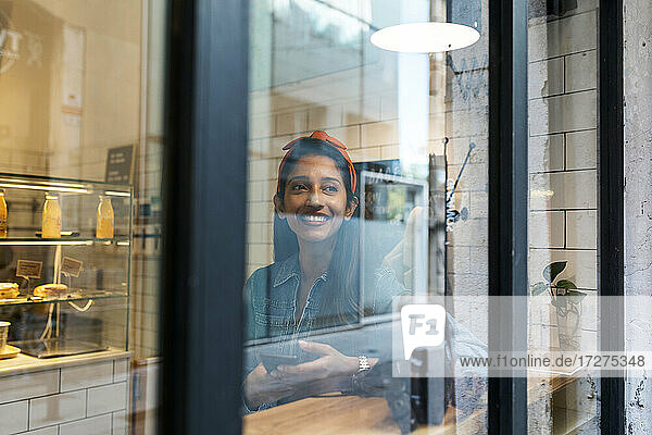 Junge Frau mit Mobiltelefon schaut durch das Fenster  während sie in einem Café sitzt