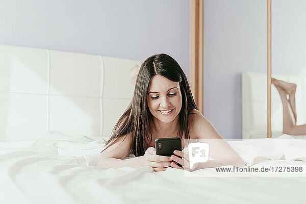 Smiling beautiful woman text messaging while lying on bed at home
