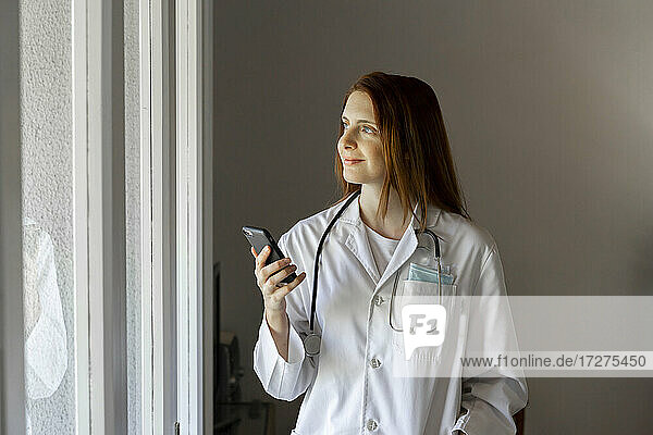 Lächelnde junge Ärztin schaut durch ein Fenster und hält ein Smartphone in der Hand