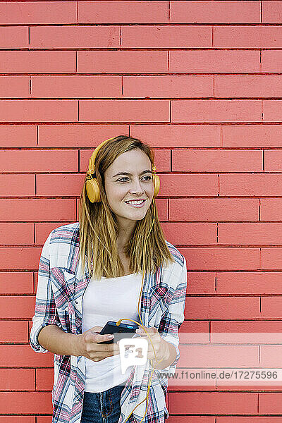 Junge Frau  die Musik über Kopfhörer hört  während sie ein Smartphone benutzt  das an einer Backsteinmauer steht