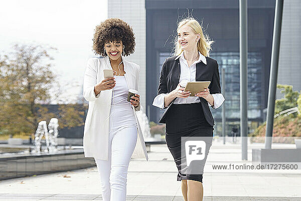 Smiling businesswomen walking with smart phone and digital tablet on footpath