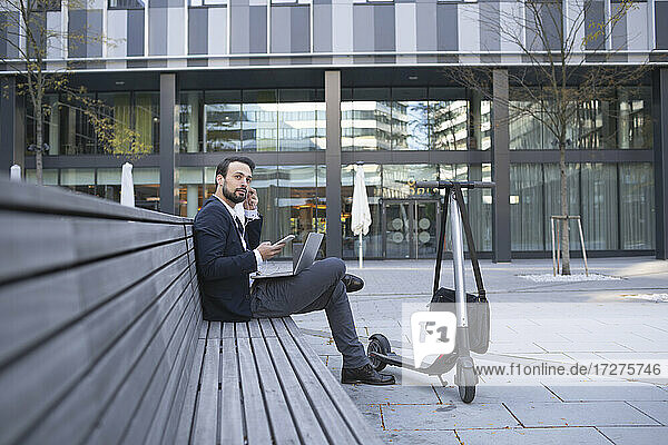 Businessman using in-ear headphones while sitting on retaining wall in city