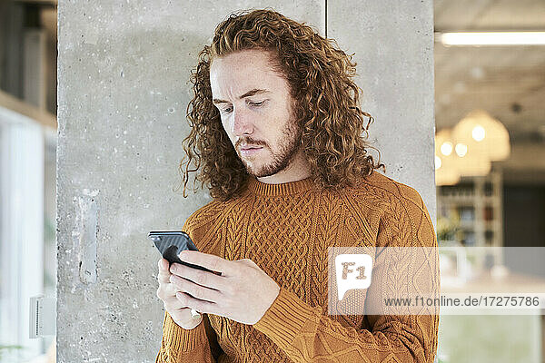 Hipster man text messaging on smart phone standing against gray column in living room at home