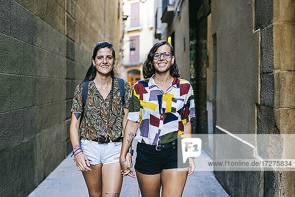 Junges lesbisches Paar  das lächelnd die Hände hält und durch eine Gasse in der Stadt geht