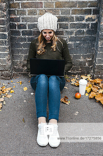 Frau  die einen Laptop benutzt  während sie auf dem Gehweg an einer Mauer sitzt