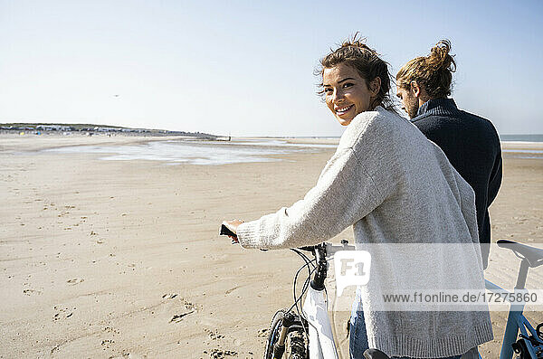 Smiling beautiful woman walking with bicycle by boyfriend while looking over shoulder at beach against clear sky on sunny day