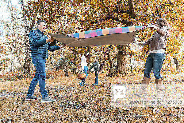Eltern legen eine Picknickdecke aus  während die Kinder im Herbst im Park stehen