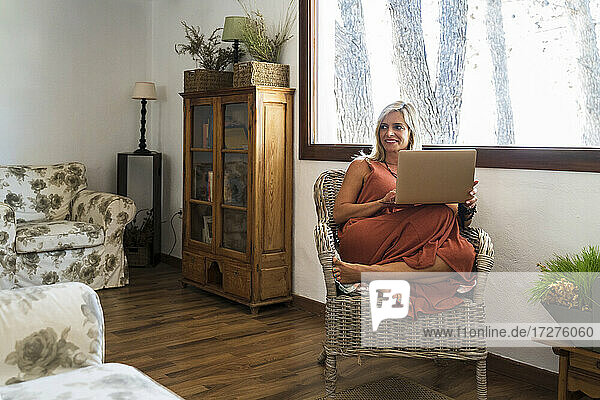 Lächelnde blonde Frau  die wegschaut  während sie mit einem Laptop auf einem Korbsessel sitzt