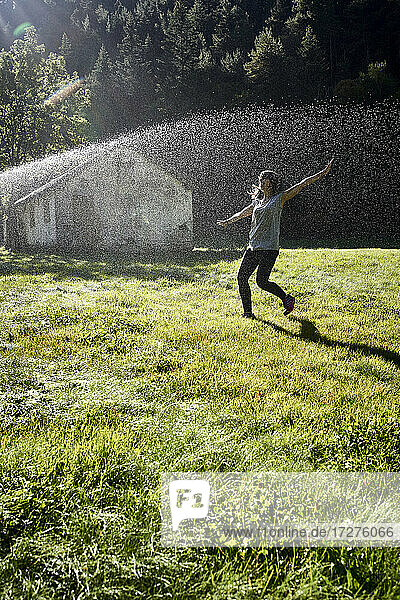 Woman standing with arms outstretched in front of sprinkler on grass during sunny day