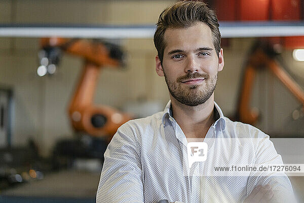 Junger Geschäftsmann lächelt  während er gegen einen Roboterarm in einer modernen Fabrik steht