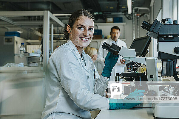 Lächelnde Frau  die einen Objektträger mit menschlichem Gehirn unter dem Mikroskop untersucht  während sie mit Wissenschaftlern im Hintergrund im Labor sitzt