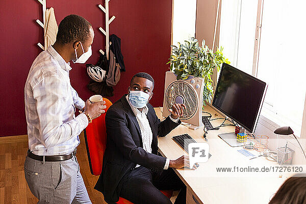 Businessmen in protective face masks discussing at desk in coworking office during COVID-19