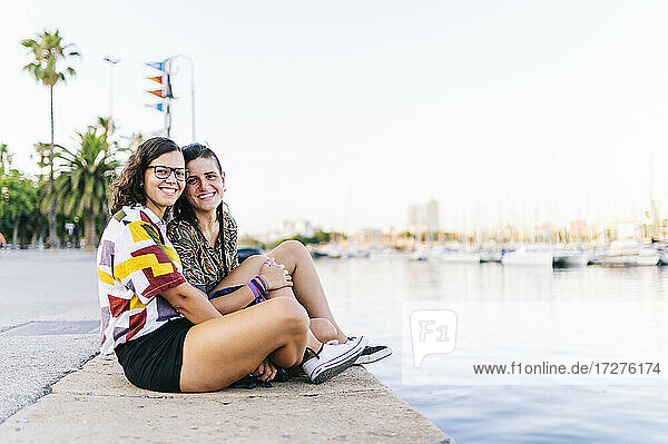 Lächelnde junge Freundinnen sitzen auf der Promenade am Hafen