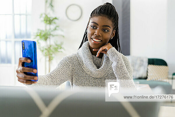 Lächelnde Studentin mit Hand am Kinn  die ein Selfie mit ihrem Smartphone macht  während sie zu Hause sitzt
