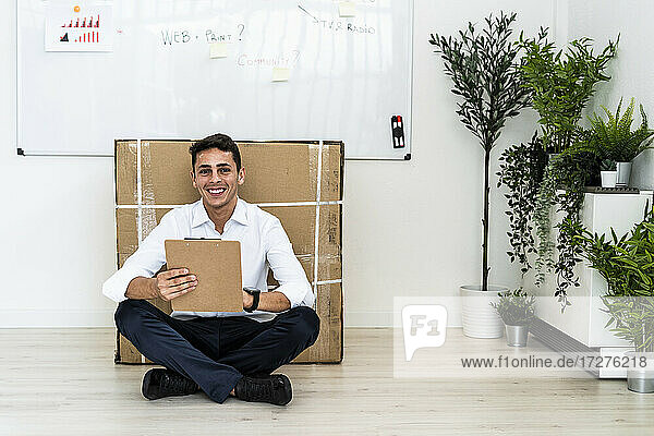Happy handsome young professional sitting with clipboard on floor against cardboard box in office