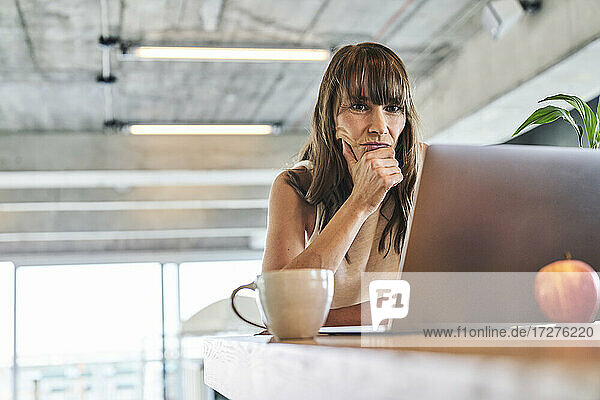 Nachdenkliche Frau mit Hand am Kinn  die einen Laptop benutzt  während sie zu Hause sitzt