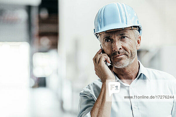 Reifer männlicher Ingenieur mit Schutzhelm  der wegschaut  während er in einer Fabrik mit einem Mobiltelefon spricht
