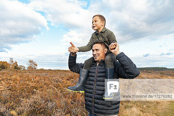 Father carrying son on shoulder standing in park against cloudy sky during autumn