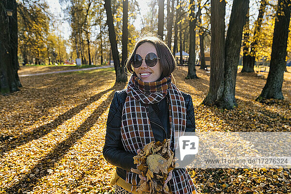 Smiling beautiful woman holding dry leaves while standing at park during autumn
