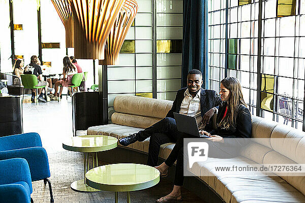 Lächelnde männliche und weibliche Geschäftsleute diskutieren über Laptop  während sie auf dem Sofa in einer modernen Bar sitzen